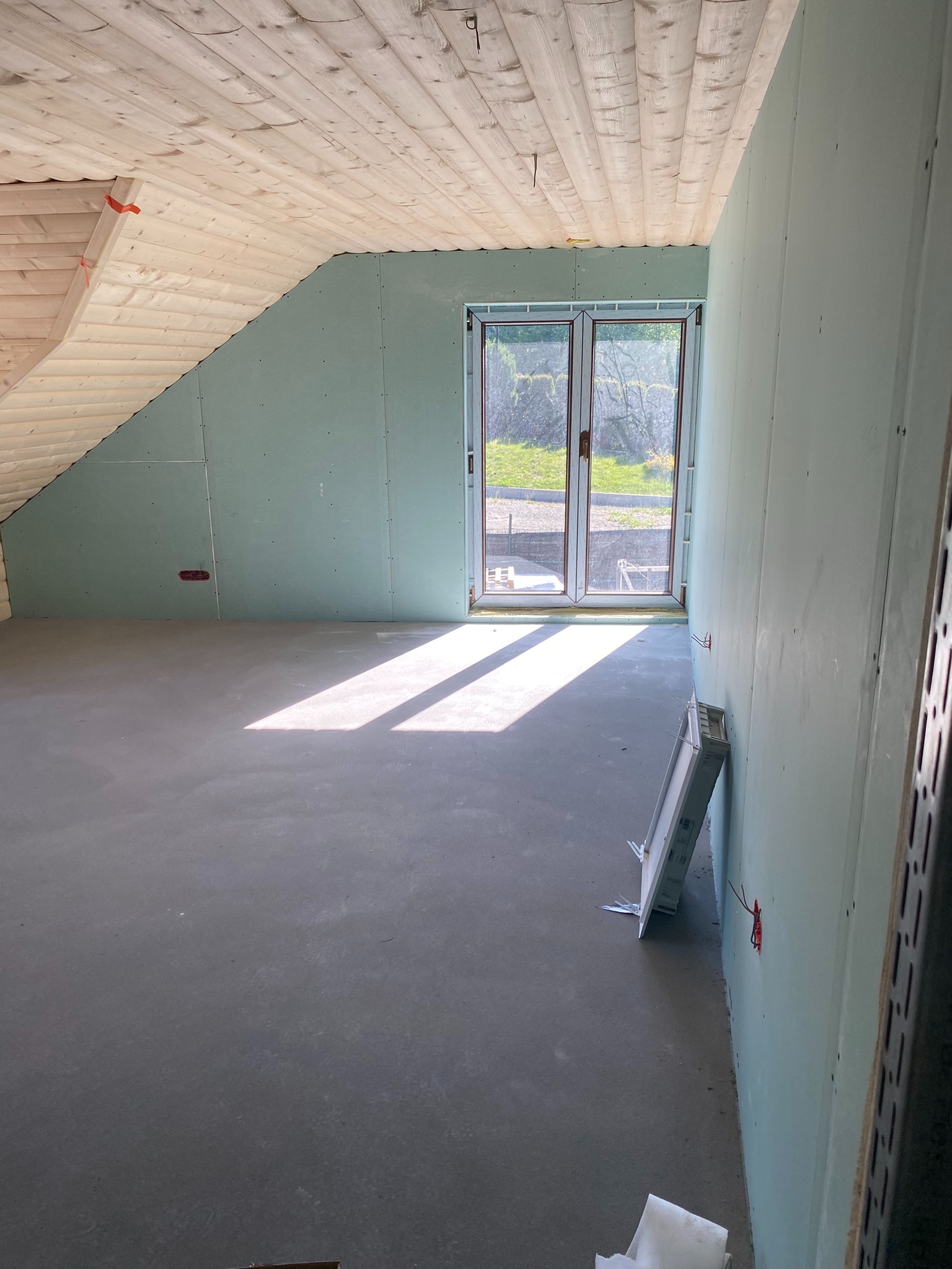 Pokój na poddaszu w trakcie wykańczania: ściany z płyt gipsowo-kartonowych, wylewka na podłodze, drewniana boazeria na suficie, okno balkonowe wpuszczające światło słoneczne.