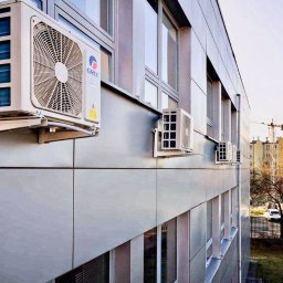 KLIMA 4U - Klima Dla Ciebie! - Zewnętrzne jednostki klimatyzacji GREE zamontowane na elewacji budynku szkoły sportowej, widoczne okna i fragment otoczenia z drzewami.