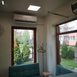 KLIMA 4U - Klima Dla Ciebie! - Klimatyzator zamontowany w biurze, widoczny przez okno ogród z tujami, w poczekalni niebieskie kanapy i wieszak na ubrania.