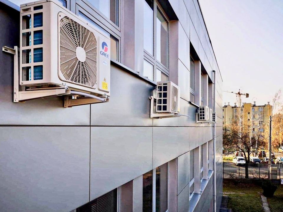Zewnętrzne jednostki klimatyzacji GREE zamontowane na elewacji budynku szkoły sportowej, widoczne okna i fragment otoczenia z drzewami.