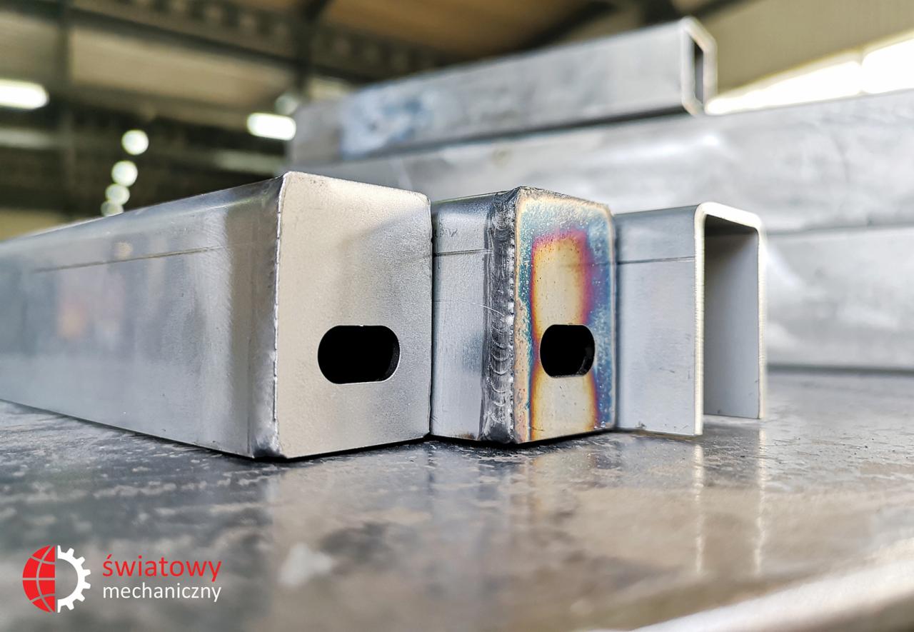 Metalowe profile o różnych kształtach, jeden z widocznymi śladami spawania i przebarwieniami termicznymi, ułożone na metalowej powierzchni.