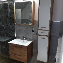 Nowoczesna łazienka we Wrocławiu po remoncie: szafka pod umywalką z drewnopodobnym frontem, wysoka szafka z białymi frontami i lustrzana szafka wisząca nad umywalką, widoczny fragment kabiny...