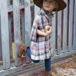Dziewczynka w brązowym kapeluszu i kraciastej pelerynie opiera nogę o szary, drewniany płot, trzymając w dłoni małe jabłko.
