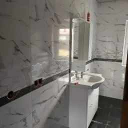 Wykończona łazienka z białymi marmurkowymi płytkami ściennymi z geometrycznym wzorem, czarnymi płytkami podłogowymi, białą szafką z umywalką i lustrem, widoczne puszki elektryczne w ścianie.