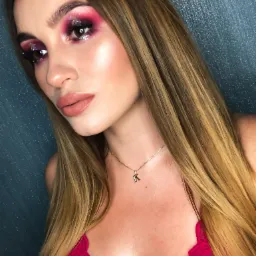 Portret kobiety z intensywnym, różowym makijażem oczu z brokatem, podkreślonymi kośćmi policzkowymi i matowymi ustami w odcieniu nude, noszącej złotą zawieszkę z literą 'K'.