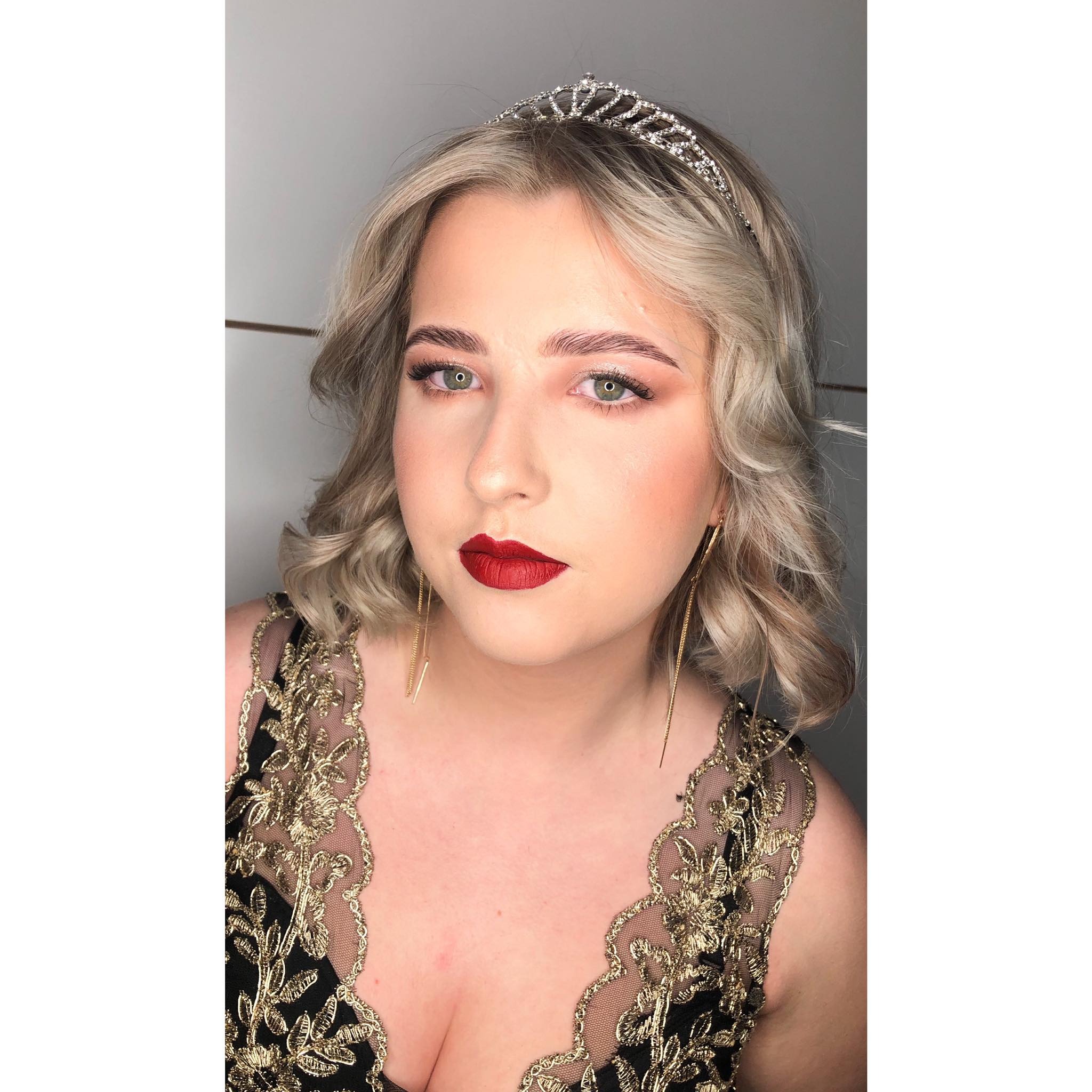 Portret kobiety z blond włosami w lokach, diademem, mocnym makijażem ust w kolorze czerwonego wina, delikatnym makijażem oczu i długimi złotymi kolczykami, ubrana w czarną sukienkę z aplikacjami...