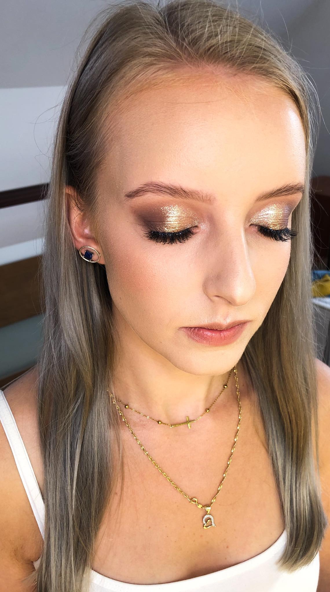 Portret kobiety z zamkniętymi oczami, prezentujący złoty, błyszczący makijaż oczu z wydłużonymi rzęsami i delikatnym konturowaniem twarzy. W uchu widoczny kolczyk z ciemnoniebieskim kamieniem. Dwa...