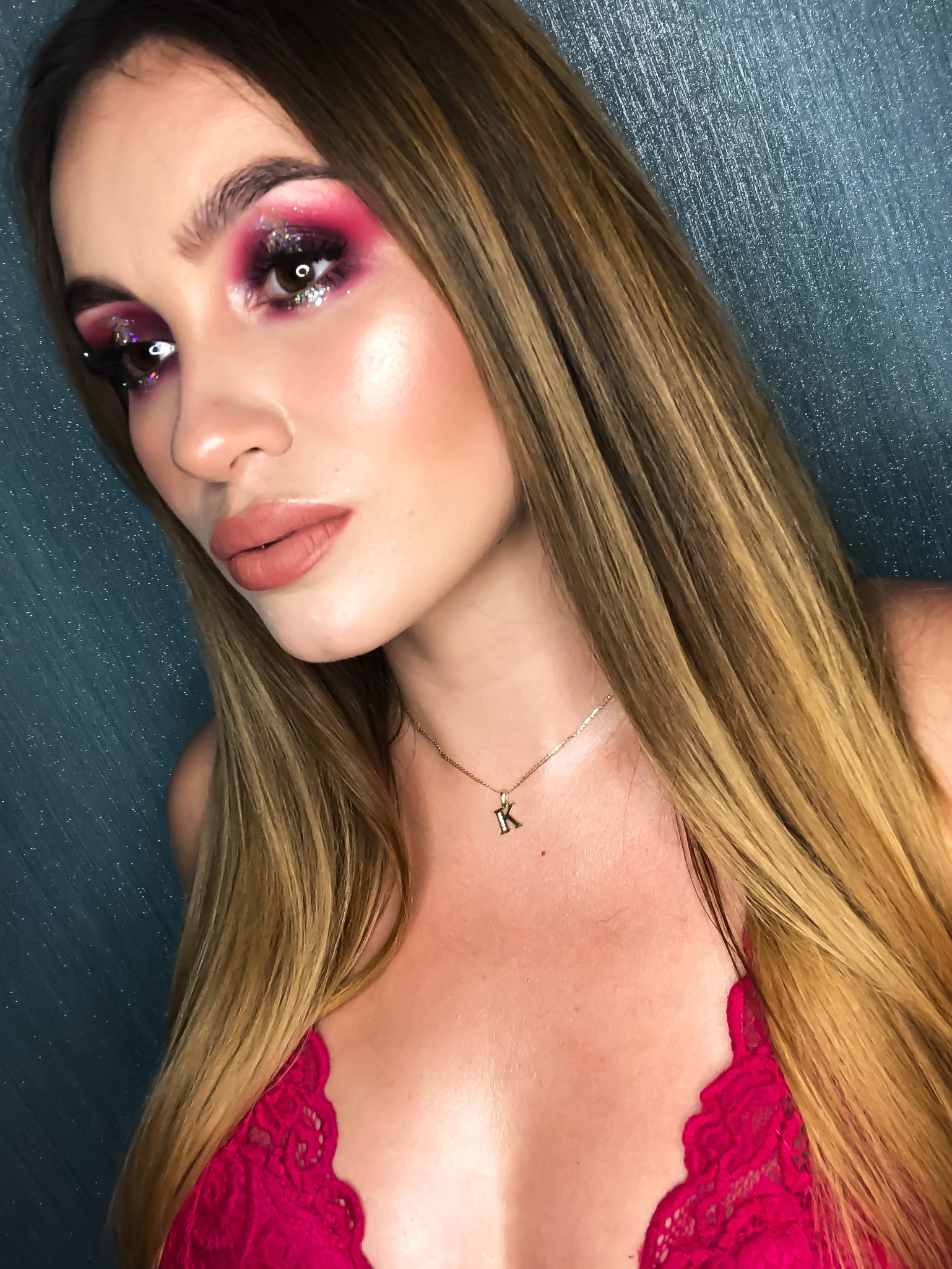 Portret kobiety z intensywnym, różowym makijażem oczu z brokatem, podkreślonymi kośćmi policzkowymi i matowymi ustami w odcieniu nude, noszącej złotą zawieszkę z literą 'K'.