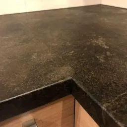 Ciemny, teksturowany blat kuchenny z widocznym narożnikiem, zamontowany na drewnianej szafce z metalowym uchwytem.