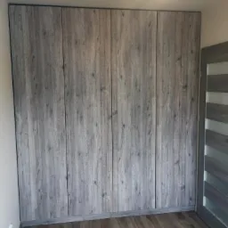 Zabudowa ściany szafą z frontami imitującymi szare drewno, widok na całą ścianę z fragmentem drzwi po prawej stronie.