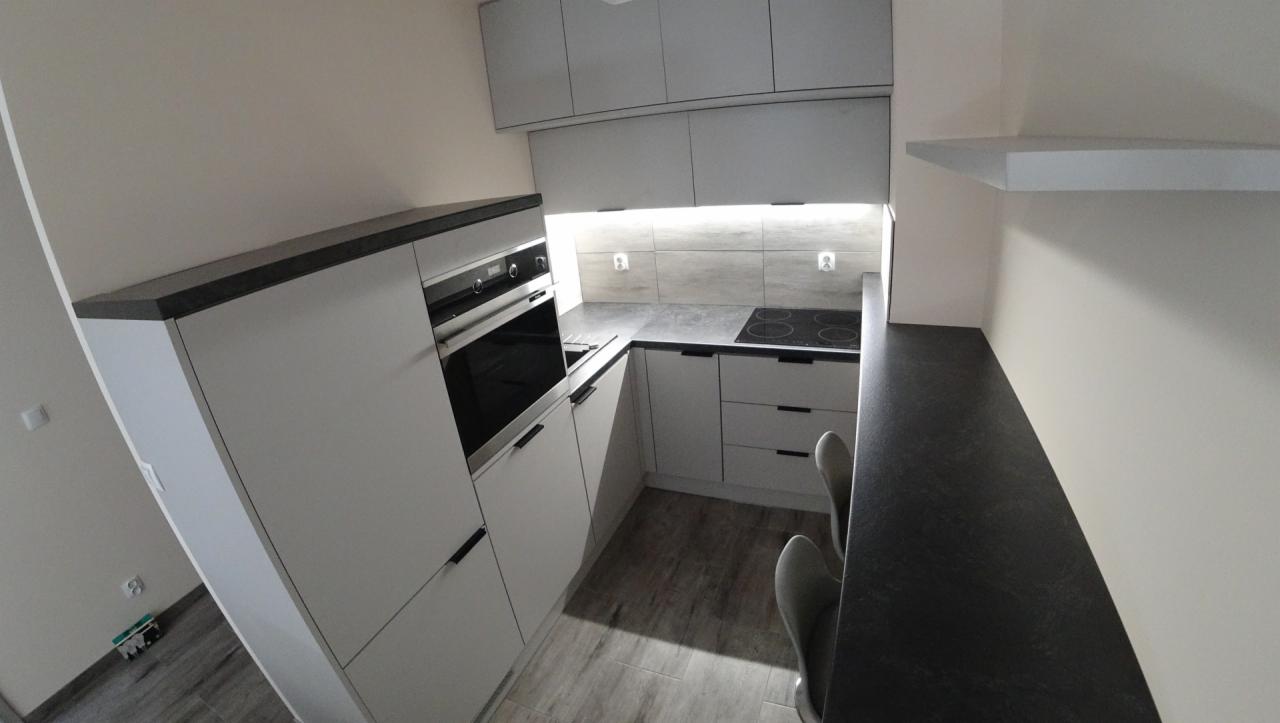 Nowoczesna kuchnia z białymi szafkami i czarnym blatem, oświetlenie podszafkowe podkreśla szary panel nad blatem, widok z perspektywy podłogi.