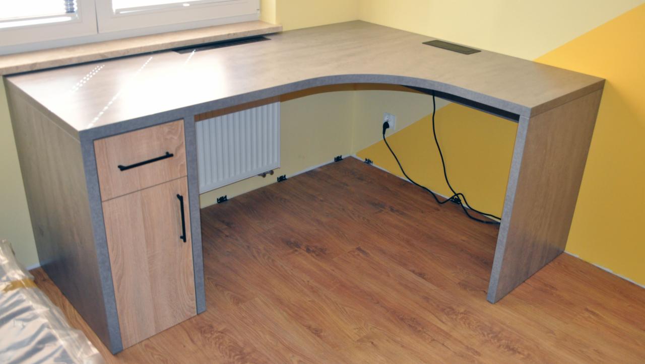 Narożne biurko z blatem imitującym beton, z drewnianą szafką z szufladą i drzwiczkami, umieszczone przy oknie i żółtej ścianie, z widocznymi kablami i fragmentem grzejnika.