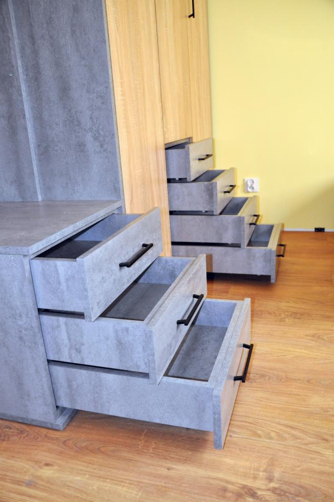Szare szafki z otwartymi szufladami i czarnymi uchwytami na tle drewnianej szafy i żółtej ściany, widoczne detale wykończenia.