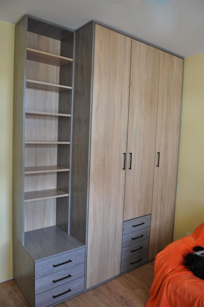 Wysoka szafa z półkami i szufladami, wykończona w odcieniach drewna i szarości, z czarnymi uchwytami, ustawiona w pokoju z żółtymi ścianami i fragmentem pomarańczowej sofy w tle.