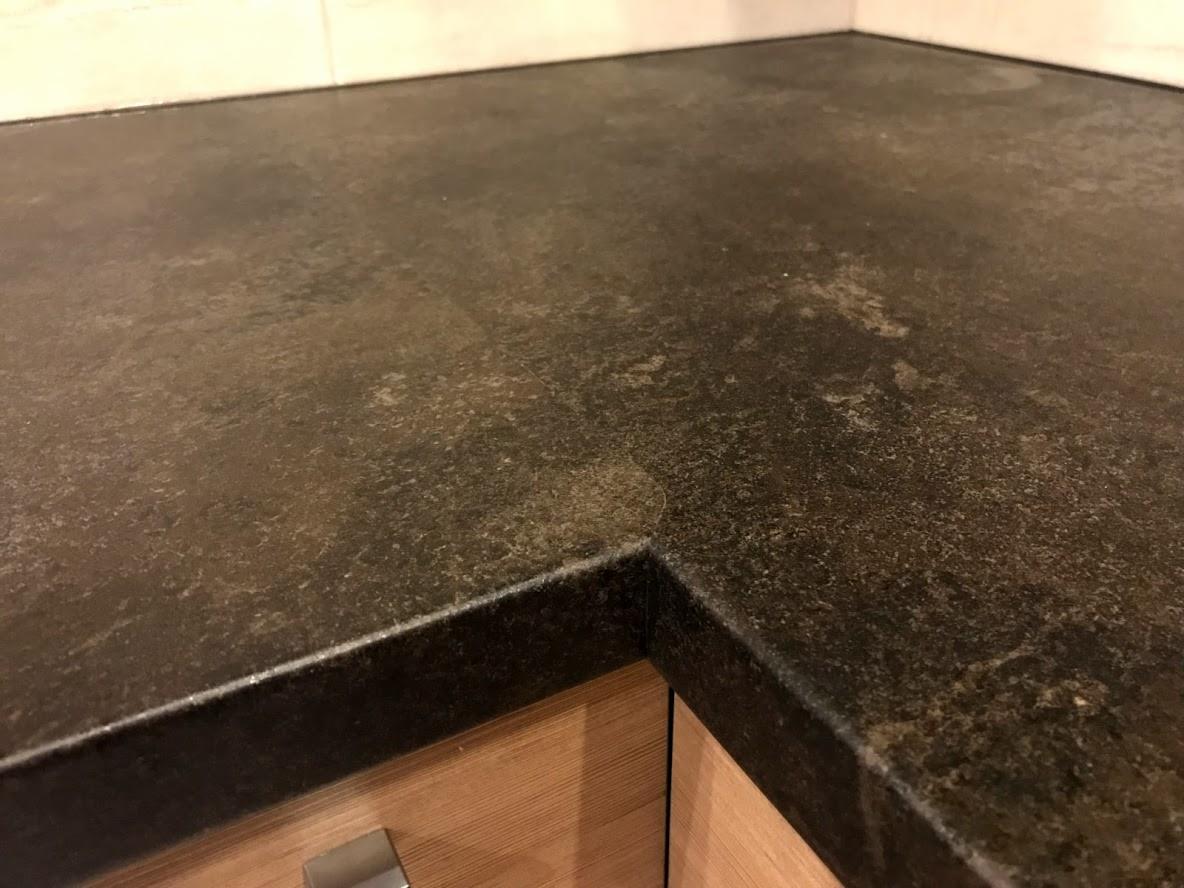 Ciemny, teksturowany blat kuchenny z widocznym narożnikiem, zamontowany na drewnianej szafce z metalowym uchwytem.