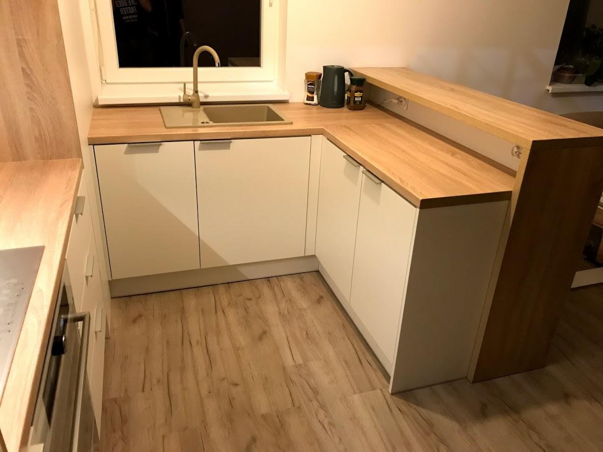 Nowoczesna kuchnia z białymi frontami szafek i drewnianym blatem, złota bateria przy zlewozmywaku, drewniana podłoga, widoczne przyprawy i czajnik.