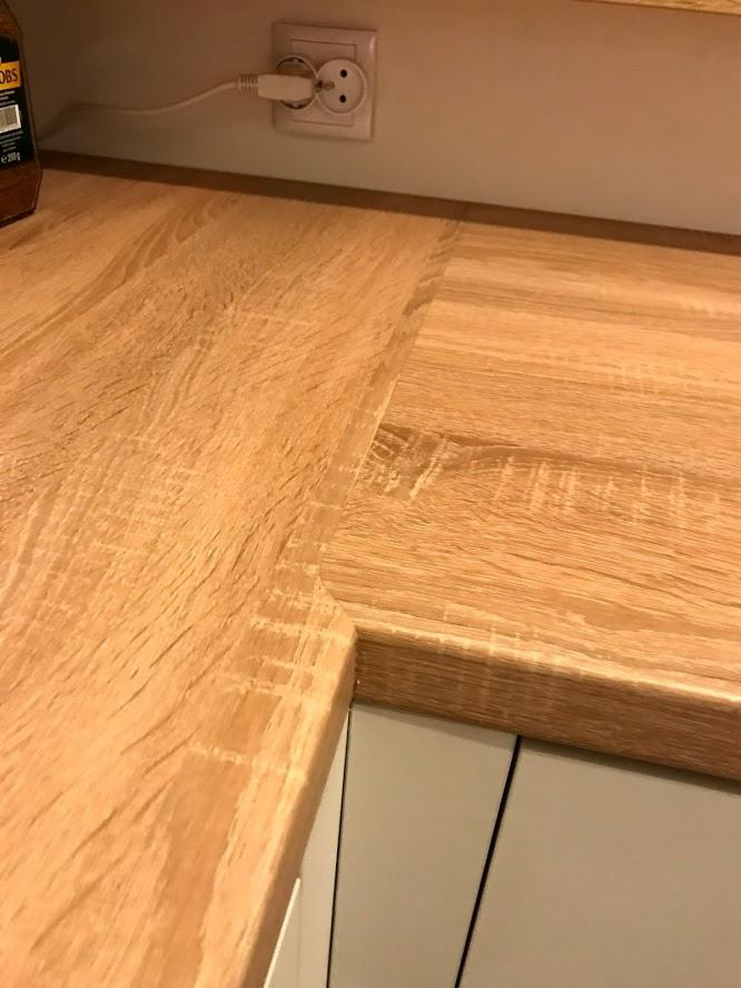 Fragment drewnianego blatu kuchennego o jasnym odcieniu dębu, z widocznym gniazdkiem elektrycznym w tle i fragmentem białych szafek pod spodem.