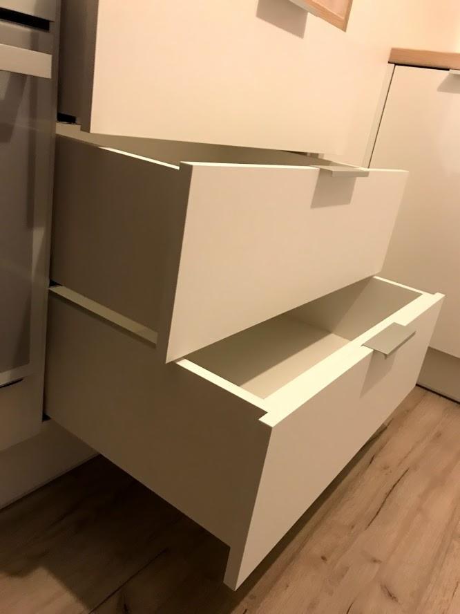 Dwie wysuwane szuflady z białymi frontami i minimalistycznymi uchwytami, zamontowane w meblu, widoczne na tle drewnianej podłogi.