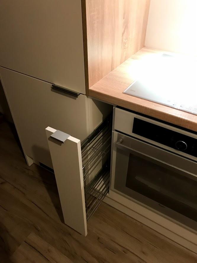 Biała kuchnia na wymiar z wysuwaną metalową szafką cargo obok piekarnika, drewniany blat i podłoga imitująca drewno.