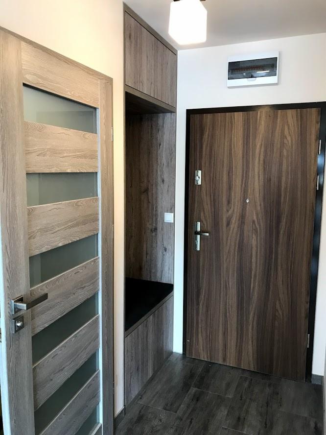 Wnętrze przedpokoju z zabudową: szafka górna, wnęka z siedziskiem, drzwi wejściowe w ciemnym fornirze i drzwi wewnętrzne z przeszkleniami w jasnym odcieniu drewna.