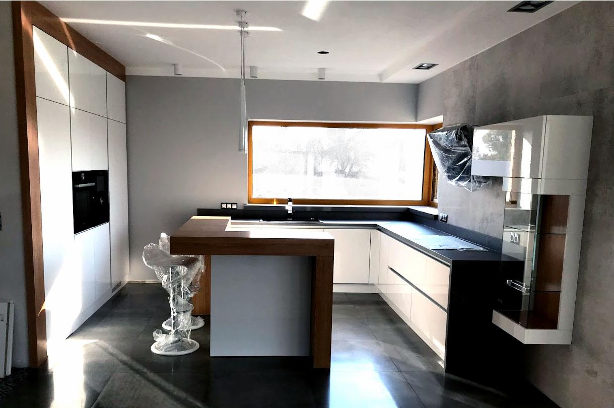 Nowoczesna kuchnia z białymi frontami, drewnianym blatem i wyspą, krzesła barowe w folii ochronnej, okno w drewnianej ramie i betonowa ściana w tle.