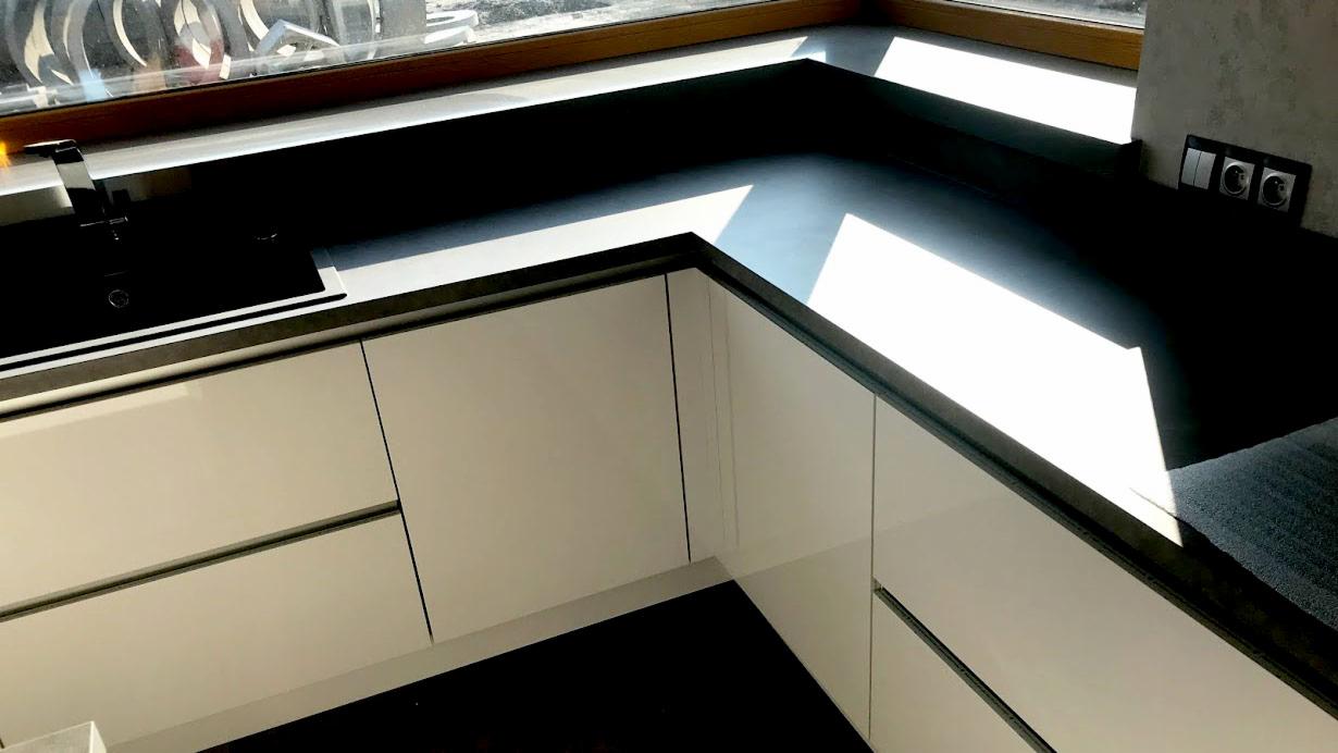 Nowoczesna kuchnia z białymi, lakierowanymi frontami szafek i ciemnym blatem, oświetlona naturalnym światłem wpadającym przez okno. Widoczny zlew i gniazdka elektryczne w ścianie.