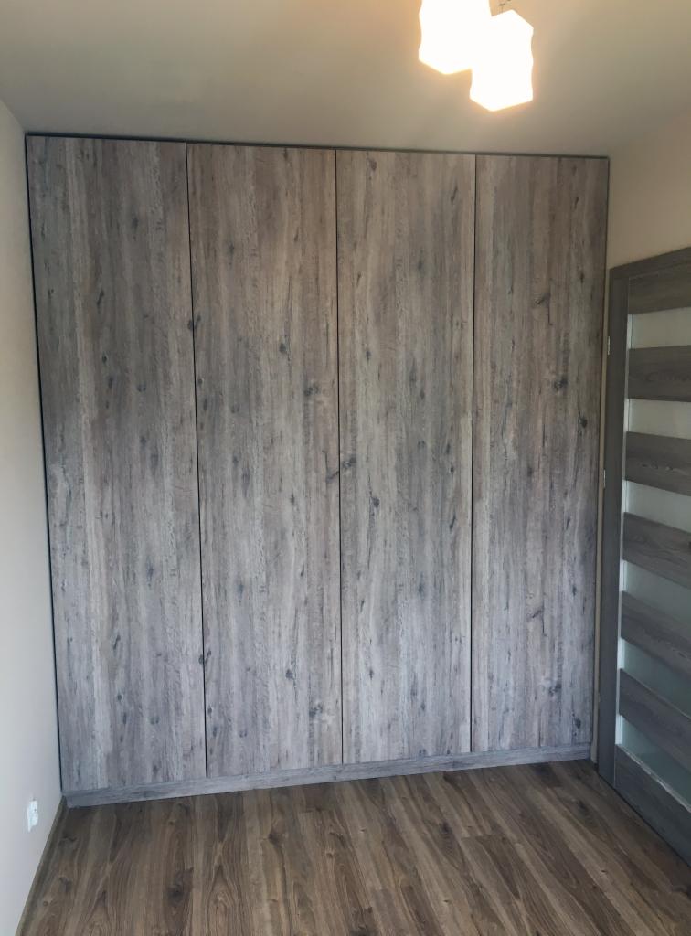 Zabudowa ściany szafą z frontami imitującymi szare drewno, widok na całą ścianę z fragmentem drzwi po prawej stronie.