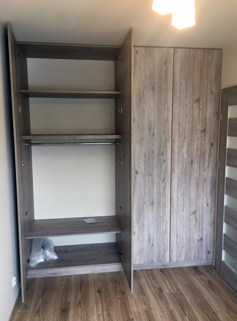 Szara szafa z otwartymi drzwiami po lewej stronie, z półkami i drążkiem na ubrania, obok zamknięta część szafy z dwojgiem drzwi o strukturze drewna, na podłodze leży folia ochronna.
