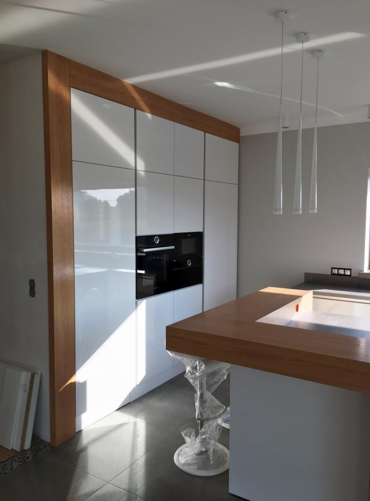 Wysoka zabudowa kuchenna z białymi, lakierowanymi frontami i drewnianą ramą, wbudowane piekarniki, wyspa kuchenna z drewnianym blatem, krzesła barowe w folii ochronnej, nowoczesne lampy wiszące.
