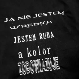 Czarna tkanina z wyhaftowanym białym napisem: 'JA NIE JESTEM WREDNA, JESTEM RUDA, a kolor ZOBOWIĄZUJE'.