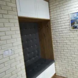 Wnęka z siedziskiem obitym ciemną tapicerką z guzikami, białe szafki wiszące i drewniane wykończenie, całość wkomponowana w ścianę z białej cegły.