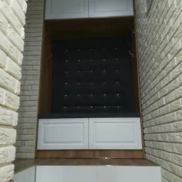 Wnęka z siedziskiem i wieszakami na ubrania, tapicerowany panel z guzikami, białe szafki górne i dolne, ściana z białej cegły.