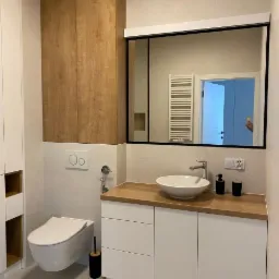 Nowoczesna łazienka z drewnianym blatem, umywalką nablatową i szafkami w kolorze białym, wisząca toaleta i wysoka szafa z drewnianymi frontami.