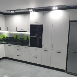 Nowoczesna kuchnia z białymi, lakierowanymi frontami, czarnymi uchwytami i blatem z motywem limonki, oświetlona punktowymi lampami sufitowymi, z wbudowanym piekarnikiem i lodówką.