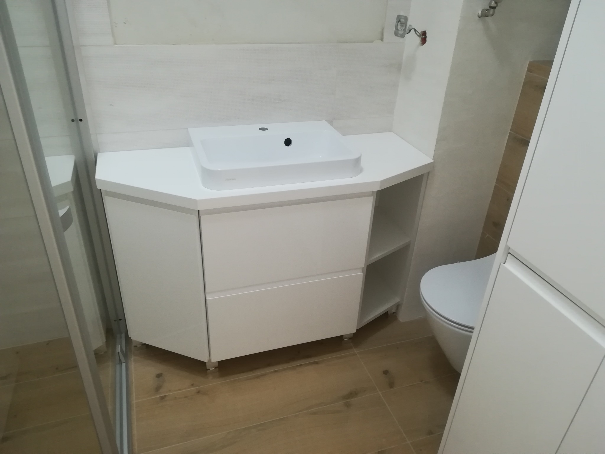 Biała, nowoczesna szafka łazienkowa pod umywalkę z dwoma szufladami i otwartą półką, stojąca na metalowych nóżkach, z umywalką nablatową, na tle płytek imitujących drewno.