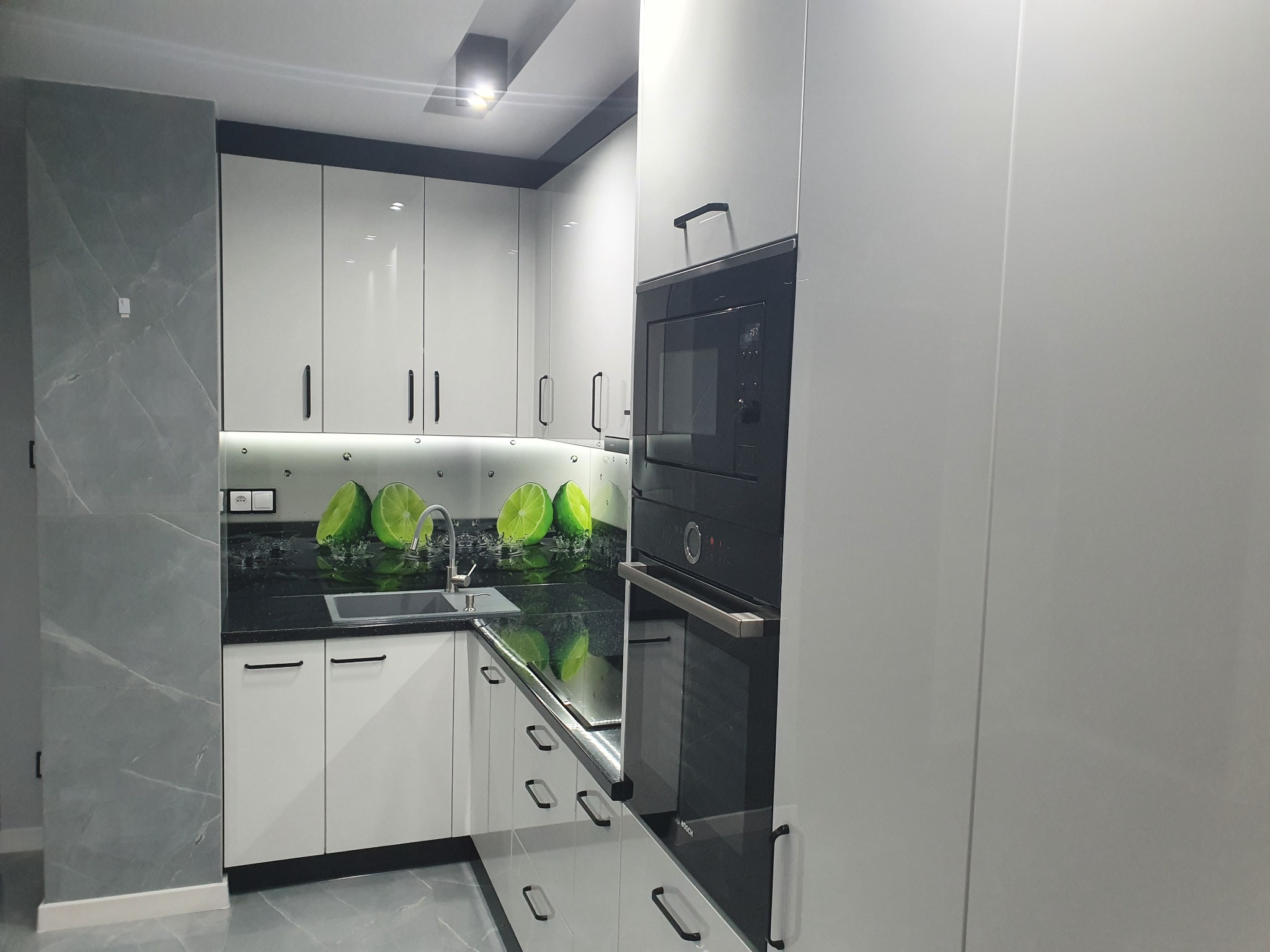 Nowoczesna kuchnia z białymi, lakierowanymi frontami, czarnym blatem z motywem limonek i wbudowanym piekarnikiem.