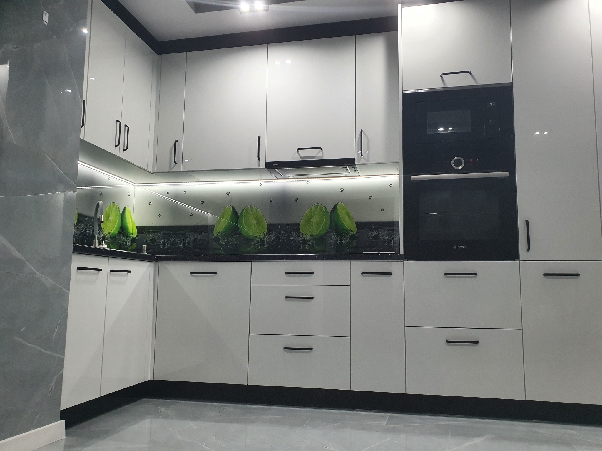 Nowoczesna kuchnia z białymi, lakierowanymi frontami, czarnymi uchwytami i panelem szklanym z motywem limonek, oświetlona listwą LED pod szafkami wiszącymi.