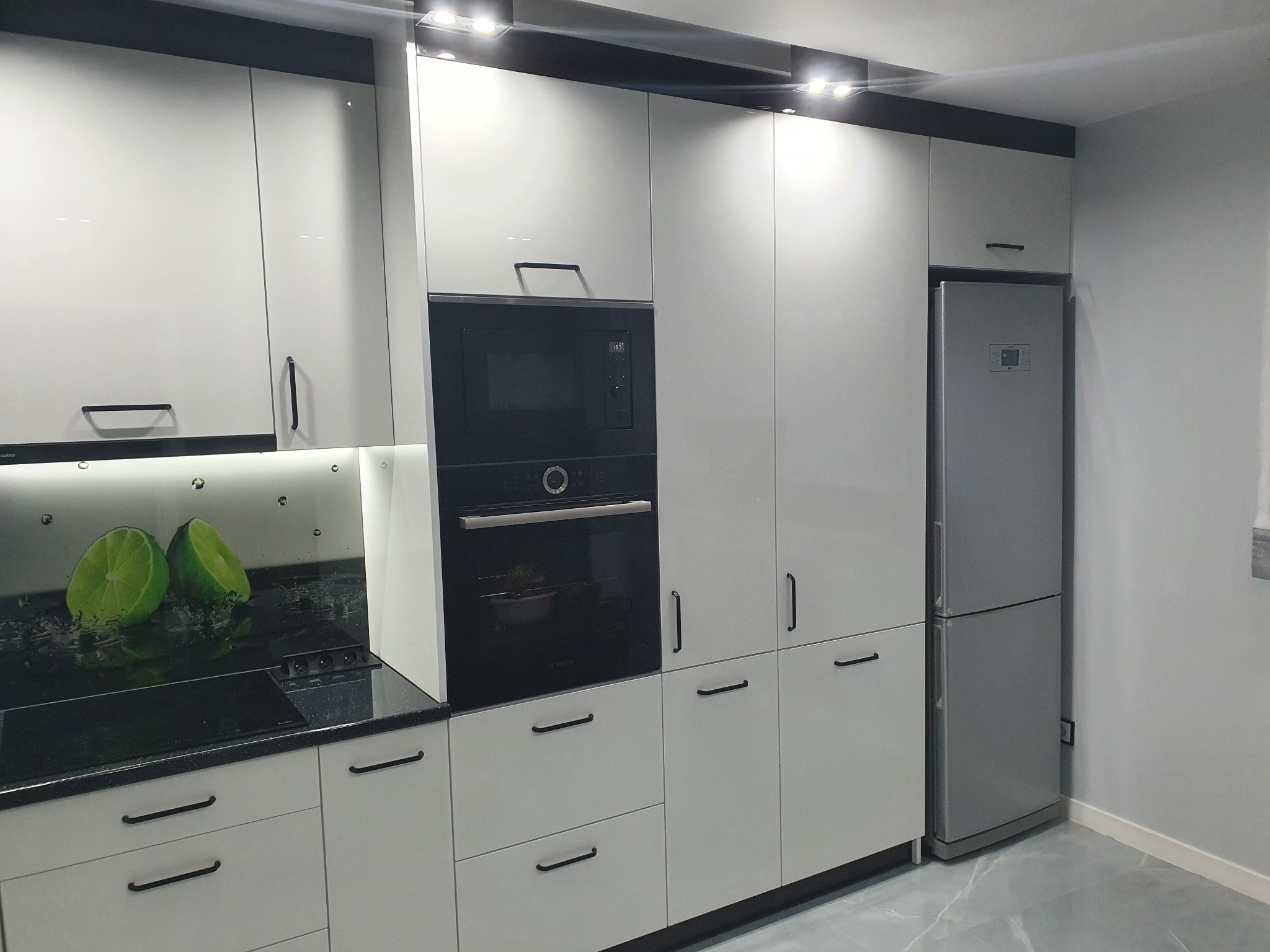 Nowoczesna kuchnia z białymi, lakierowanymi frontami, czarnymi uchwytami i blatem z motywem limonek na ścianie, zabudowana z piekarnikiem, mikrofalówką i lodówką.