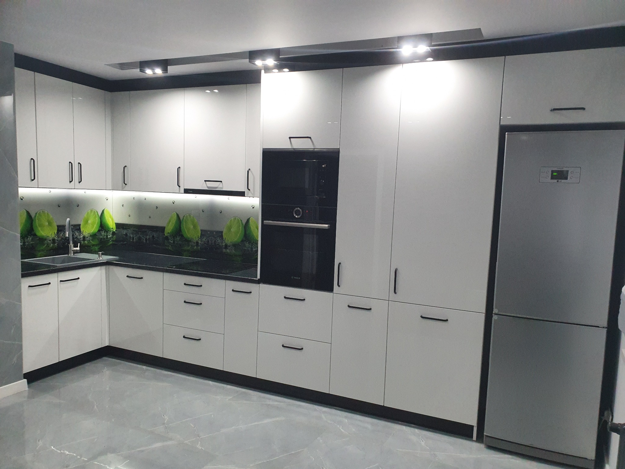 Nowoczesna kuchnia z białymi, lakierowanymi frontami, czarnymi uchwytami i blatem z motywem limonki, oświetlona punktowymi lampami sufitowymi, z wbudowanym piekarnikiem i lodówką.
