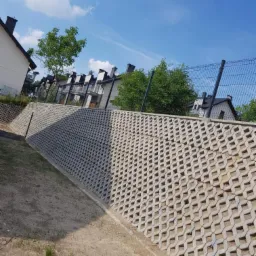 Ściana oporowa z ażurowych betonowych elementów, zabezpieczająca skarpę przy nowym osiedlu mieszkaniowym, widoczna siatka ogrodzeniowa i fragmenty budynków.