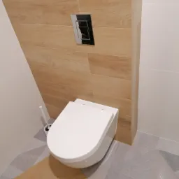 Nowoczesna toaleta podwieszana z drewnopodobną obudową stelaża i chromowanym przyciskiem spłukującym, na podłodze geometryczne płytki.