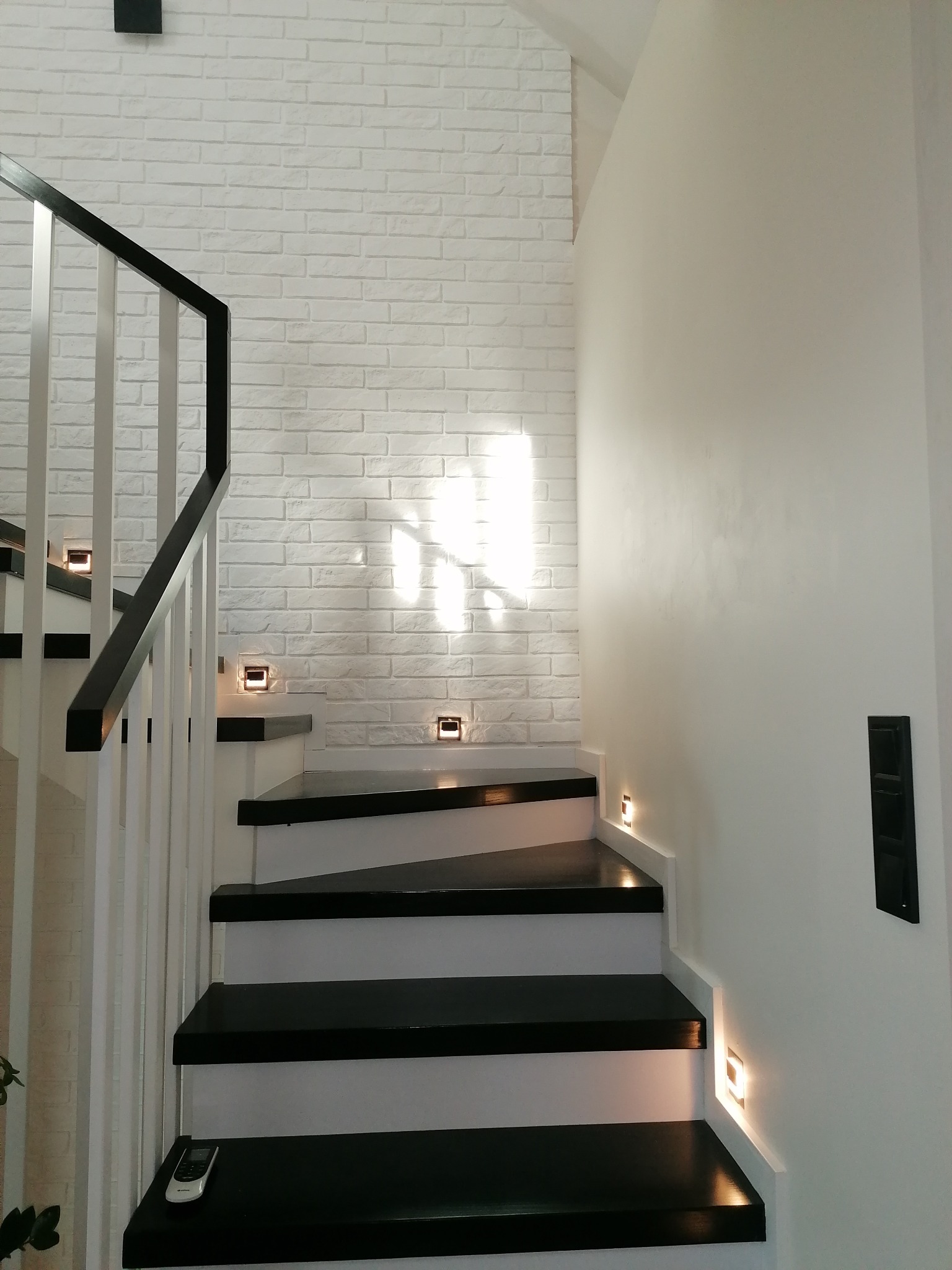 Nowoczesne schody z czarnymi stopniami i białymi podstopnicami, oświetlone punktowymi lampami w ścianie, obok ściana wykończona białą cegłą.
