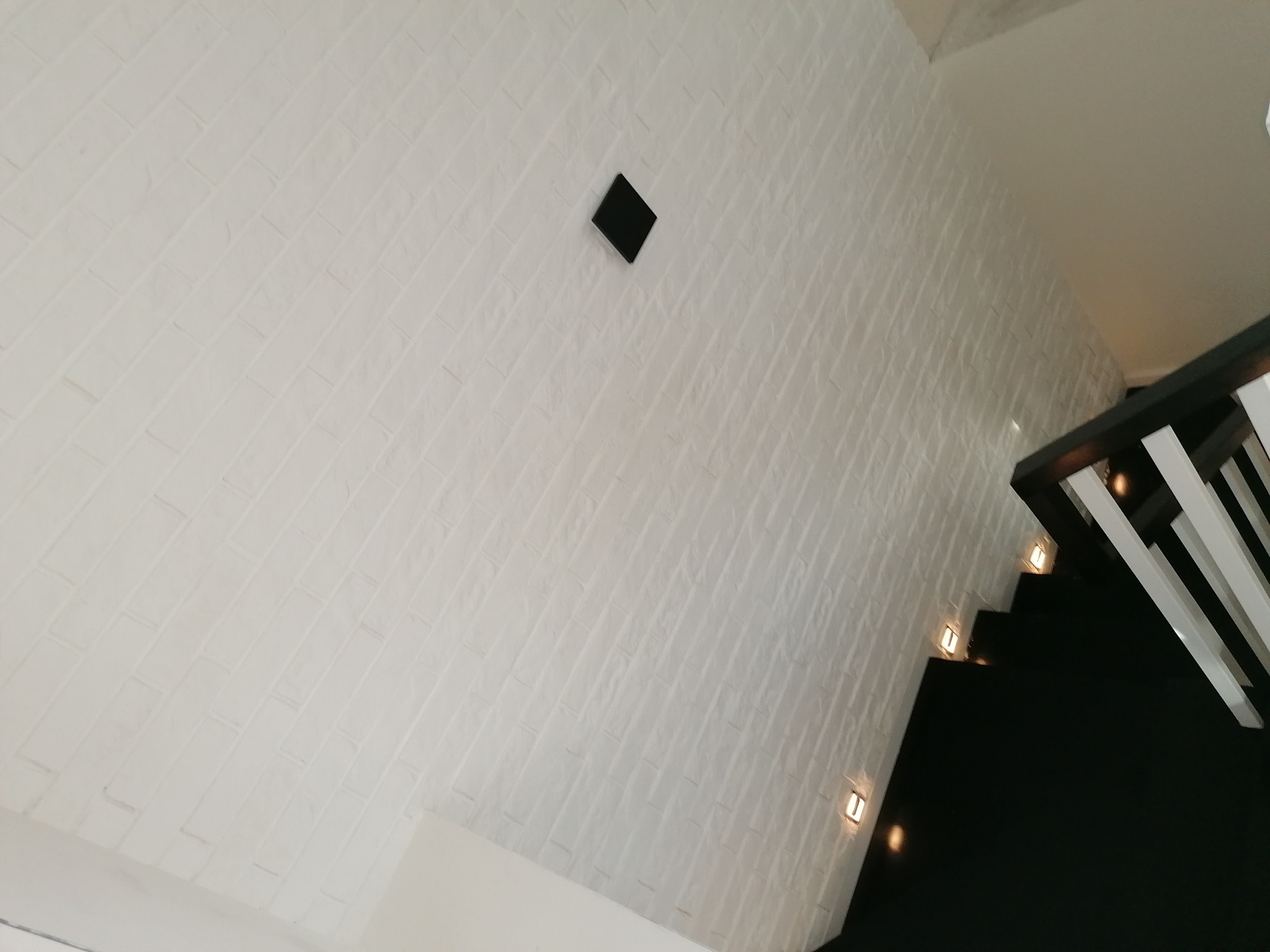 Biała ściana z dekoracyjną fakturą cegiełek, oświetlona schodami z czarnymi stopniami i białymi barierkami, dodatkowo w ścianie zamontowany czarny element.