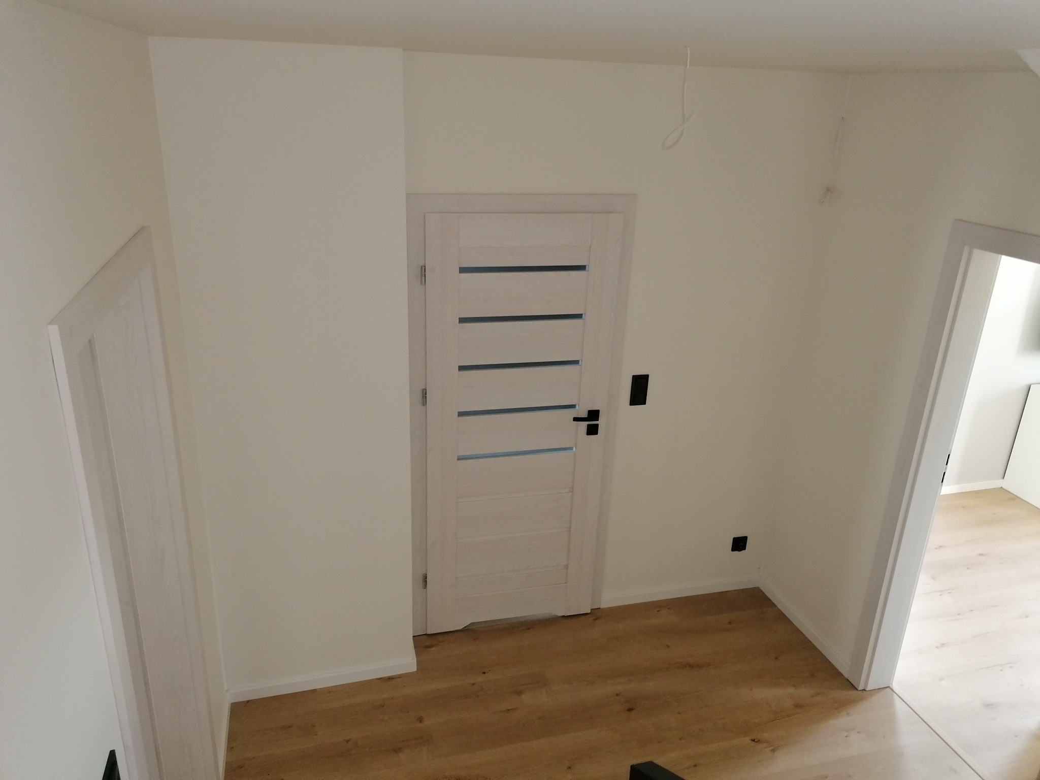 Korytarz po remoncie z dwiema białymi ościeżnicami, drewnianą podłogą i pomalowanymi na biało ścianami; widoczne wystające ze ściany kable