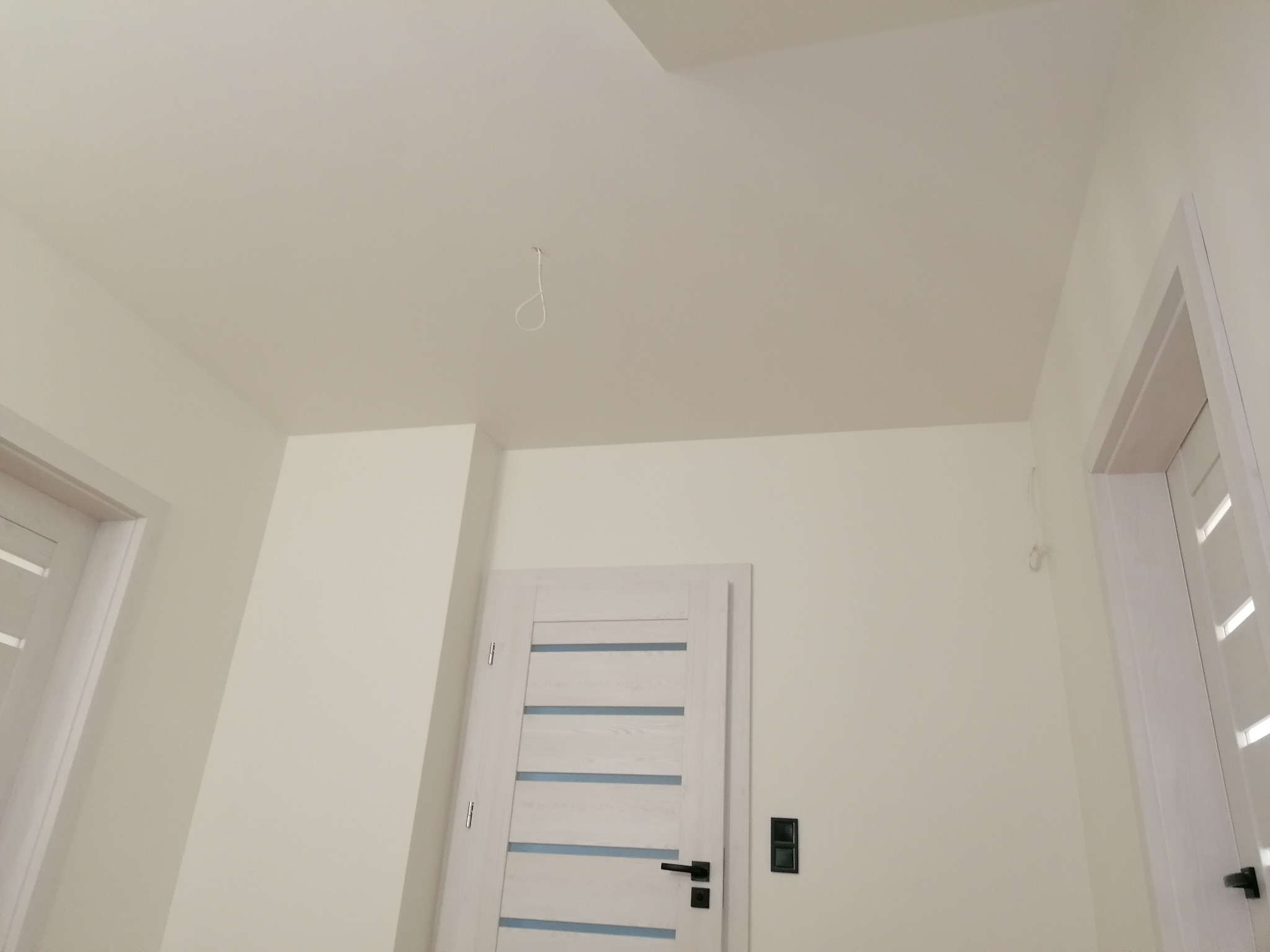 Wnętrze po malowaniu na biało, widoczna gładka powierzchnia ścian i sufitu, białe drzwi z poziomymi przeszkleniami, wystający kabel oświetleniowy z sufitu.