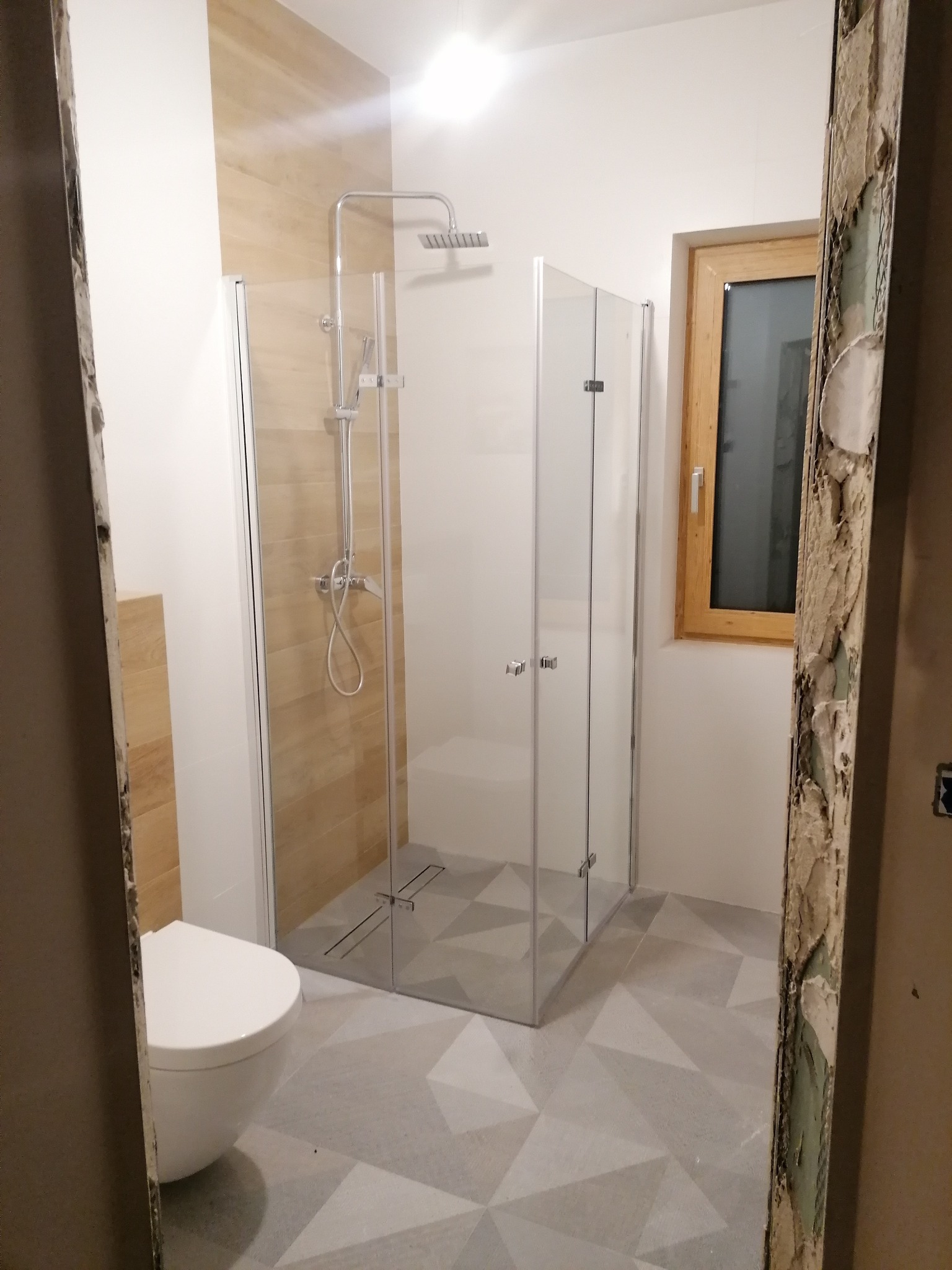 Nowoczesna łazienka po remoncie z kabiną prysznicową typu walk-in, wiszącą toaletą i geometryczną podłogą, widoczna przez futrynę w stanie surowym.