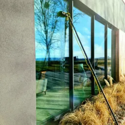 Czyszczenie okien nowoczesnego domu za pomocą teleskopowego kija z myjką, widoczne smugi wody na szybie, w tle odbicie drzew i błękitne niebo, ozdobne trawy przy elewacji.