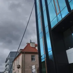 Mycie szklanej elewacji nowoczesnego biurowca przy użyciu lancy teleskopowej z ziemi; w tle budynek mieszkalny i pochmurne niebo.