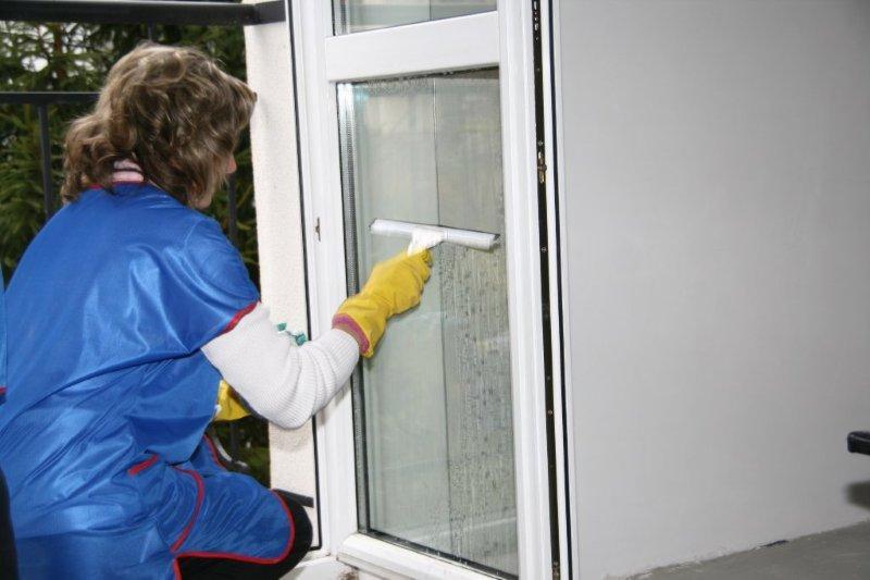 Osoba w niebieskim fartuchu i żółtych rękawicach czyści okno za pomocą ściągaczki, widoczne smugi wody na szybie.