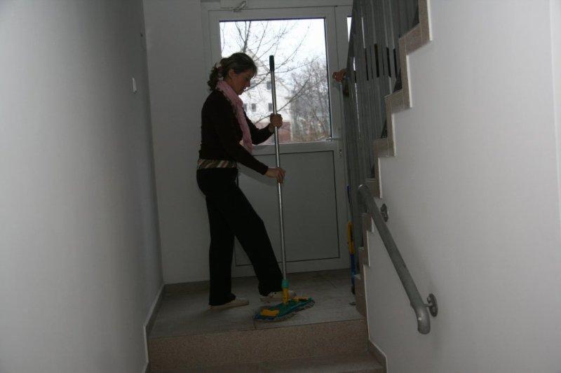 Kobieta w ciemnym stroju myje podłogę mopem na klatce schodowej wewnątrz budynku, widoczne schody z metalową barierką i okno w tle.